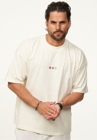 Cremfarbener oversized T-Shirt aus weicher Baumwolle, mit einem kleinen mehrfarbigen Grafikdesign auf der Brust. Klassischer Rundhalsausschnitt mit kurzen Ärmeln.