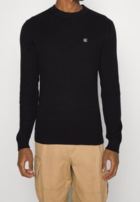 Svart ribbad tröja med rund halsringning och en liten logotyppatch på bröstet. Har långa ärmar och en figurnära design.