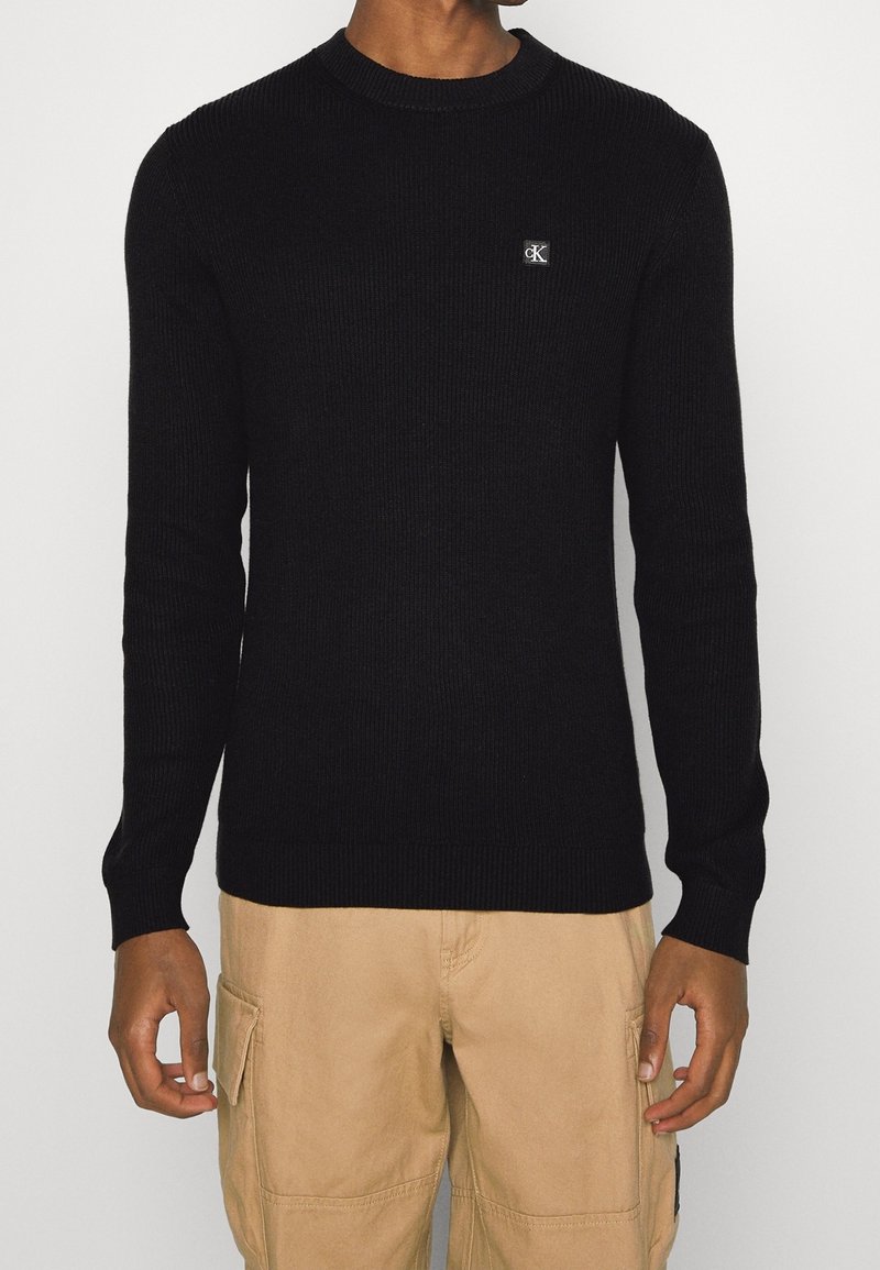 Svart ribbad tröja med rund halsringning och en liten logotyppatch på bröstet. Har långa ärmar och en figurnära design.