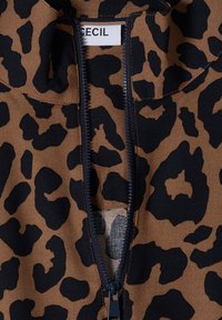 Tissu imprimé léopard marron et noir avec une fermeture éclair noire partiellement ouverte révélant une étiquette blanche avec un texte noir "CECIL" à l'intérieur.