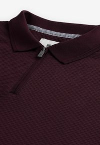 Polo de culoare vișinie, dintr-un material texturat, cu guler scurt și fermoar frontal. Gulerul interior are un model în carouri.
