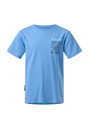 Chłopiec stoi z rękami w kieszeniach, ubrany w szarą bluzę na suwak, niebieski t-shirt z nadrukiem, niebieskie dżinsy i białe trampki z czarnymi paskami.
