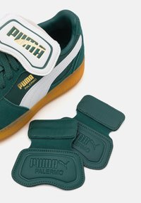 Zielone zamszowe sneakersy z białymi akcentami i gumową podeszwą, z dużym białym paskiem z złotym napisem. W zestawie znajdują się dwa turkusowe skórzane wstawki.