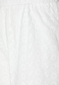 Tissu blanc brodé avec des motifs floraux, présentant une surface texturée et une taille élastique froncée, adapté pour un usage décontracté.