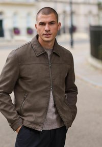 Brown leather zip-up jacket with a collared neckline, side pockets, and a smooth texture, layered over a light sweater.