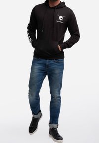 Felpa nera con cappuccio con grafica di teschio, maniche lunghe e tasca frontale. Indossata con jeans denim blu e scarpe nere, che mostrano un look casual.