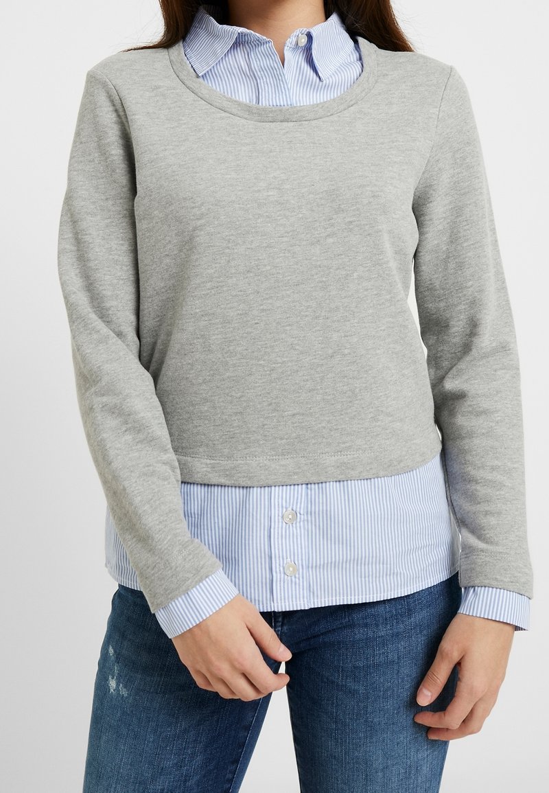 Sweat-shirt gris à manches longues avec un col rond, porté par-dessus une chemise à col bleu clair et blanc à rayures. Jean en denim visible en dessous.