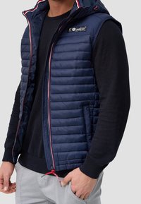 Gilet imbottito blu navy con cappuccio, texture trapuntata, chiusura frontale con cerniera, dettagli in rosso e toppa con logo; indossato sopra un maglione nero.