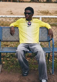 Nike dzeltena garām piedurknēm krekls ar vertikālajām svītrām, pelēki platās bikses, melnas kurpes un saulesbrilles, sēž uz zilā metāla soliņa.