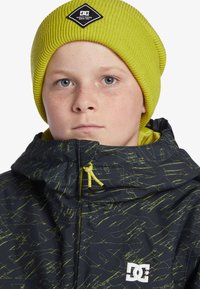 Svart jacka med gröna abstrakta mönster, justerbar huva och gul dragningssnodd. Matchas med en ribbad gul mössa med en logopatch.