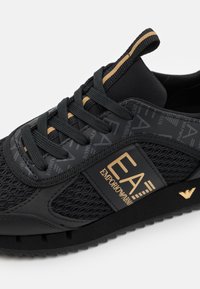Zapatilla negra que presenta materiales de malla y lisos, acentuada con detalles y logotipos dorados, puntera redonda y suela negra texturizada.