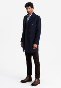 Cappotto navy doppiopetto in misto lana, con grandi bottoni, rever a punta, tasche laterali, indossato con pantaloni attillati marroni e stivali in pelle.