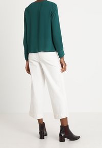 Blusa de manga larga en verde azulado con un dobladillo trasero redondeado, combinada con pantalones cortos de pierna ancha blancos y botas tobilleras negras con un acabado texturizado.