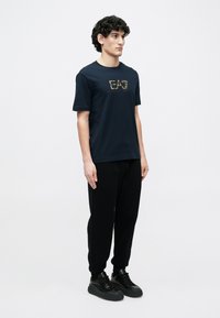 Tengerészkék pamut póló arany "EAT" felirattal, fekete melegítőnadrággal és fekete sportcipővel párosítva. Egyszerű, laza szabás, minták nélkül.