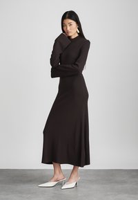 Long, fitted dark brown dress with high neckline and long sleeves, featuring a side slit and smooth, stretchy fabric. Paired with white mules.