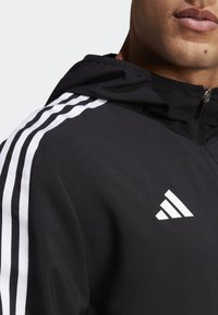 Černá sportovní bunda s kapsou na zip, kapucí a třemi bílými pruhy na rameni. Logo umístěné výrazně na hrudi.