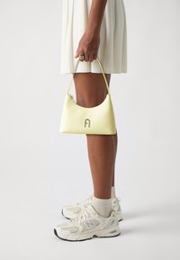 Gelbe Lederhandtasche mit geschwungener Form und silbernem Logo. Der Griff ist dünn. Modell trägt ein weißes Kleid und cremefarbene Sneaker.