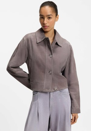 Kvinde iført en cropped, knaplukket, grå ruskindsjakke og højttaljede, lysegrå, vide bukser, poserer med én hånd bag ryggen.