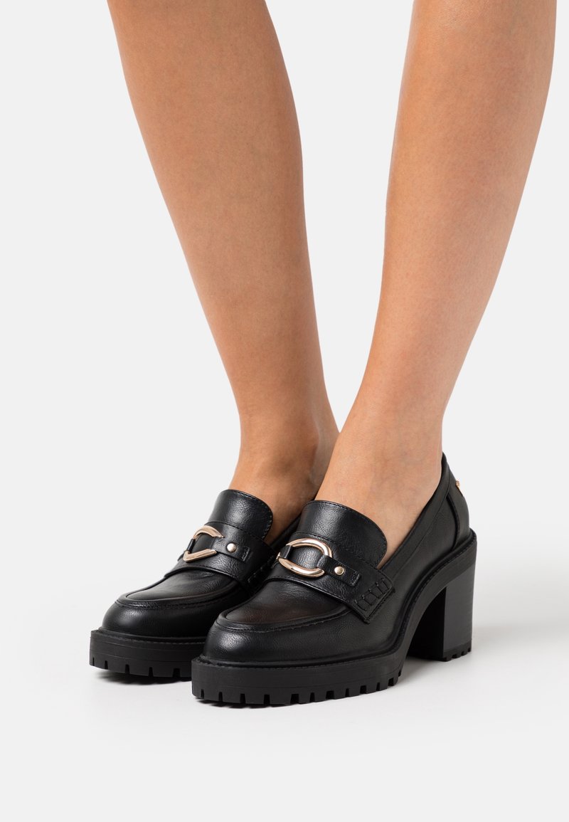 Mocassins en cuir noir à talon épais avec détail de boucle dorée portés sur une paire de jambes nues sur un fond uni.