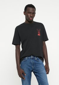 Homem vestindo uma t-shirt preta com um gráfico de coelho vermelho e jeans azuis, de pé contra um fundo branco simples.