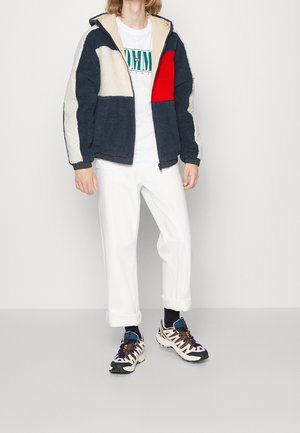 Giacca in pile di colore blu navy e crema con pannelli rossi e bianchi, chiusura a zip, polsi elasticizzati, abbinata a una t-shirt bianca e pantaloni bianchi larghi.