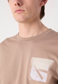T-shirt en coton brown clair avec un col rond et un design de poche imprimée à l'avant présentant des formes abstraites dans des couleurs douces.