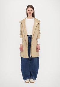 Giacca beige con cappuccio e chiusura con zip, abbinata a jeans in denim blu larghi. Il modello indossa una maglietta bianca a maniche lunghe sotto.
