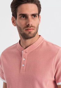 Růžová polo košile s texturovanou tkaninou, má límec a zapínání na tři knoflíky, krátké rukávy s kontrastními bílými akcenty.