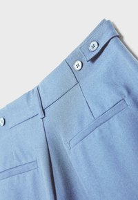 Pantalon bleu clair en gros plan montrant la taille avec deux boutons et passants de ceinture, ainsi que les poches arrière horizontales sur un fond uni.