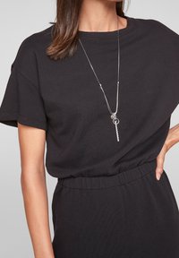 Camiseta negra de manga corta con escote redondo, combinada con un collar de pendant de plata que presenta un diseño de llave, usado sobre una falda negra a juego.