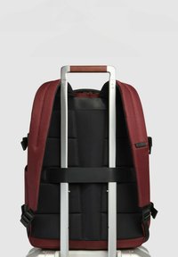 Rode en zwarte rugzak met gewatteerde achterkant, verstelbare banden en een opgeborgen trolleysgreep. Zichtbare textuur en gedetailleerde naden.