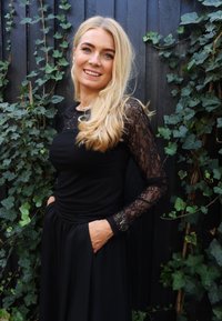 Must kleit pikkade pitsist varrukate ja vormitud korsetiosaga. Seelik on voogav. Modell seisab puidust tausta ees, millel on viinapuu.