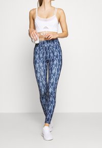 Bílé sportovní podprsenka s síťovými detaily, v kombinaci s těsnými modrými legínami se abstraktním vzorem. Model nosí bílé tenisky.