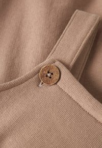 Tkanina v bež barvi z teksturirano površino, s lesenim gumbom s cvetličnim motivom, ki krasi naramnico. Čiste linije in preprost dizajn.