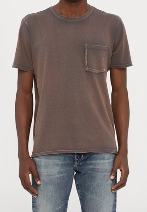 Mann trägt ein braunes kurzärmeliges T-Shirt mit Brusttasche und blaue ausgewaschene Jeans, steht vor einfachem Hintergrund.