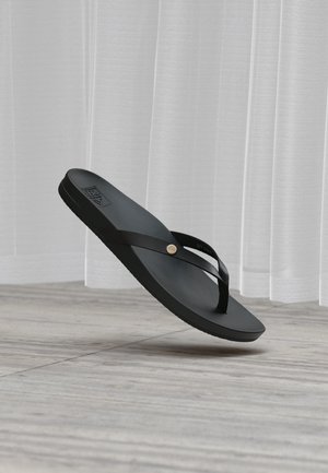 Sandale flip-flop en caoutchouc noir avec des fines bretelles et un petit détail métallique rond, flottant au-dessus d’un sol en bois devant des rideaux blancs.