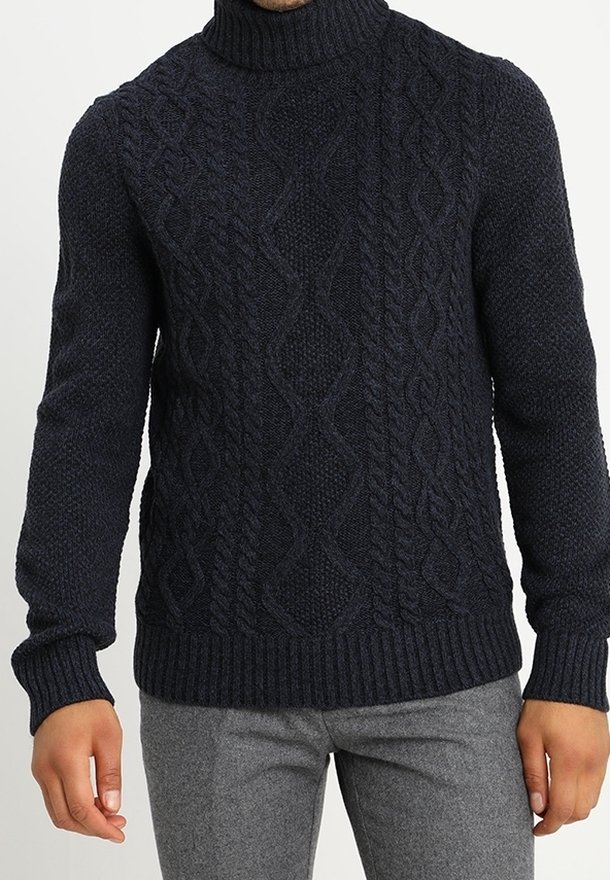 Homme portant un pull à col roulé en laine torsadée bleu marine foncé et un pantalon gris clair, debout, les bras détendus le long du corps.