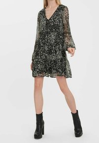 Robe fleurie noire à manches longues transparentes, encolure en V, volants à étages et tissu léger texturé. Associée à des bottes à talons noires.