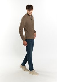 Beige kötött, zipzáros pulóver bordás mandzsettával és aljjal, kék farmerrel és világos színű sportcipővel párosítva, modell fehér háttér előtt sétál.