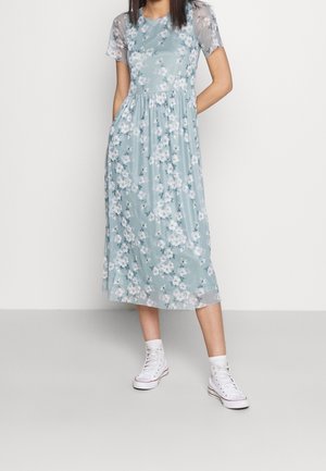 Kwiatowa sukienka midi w jasnoniebieskim kolorze z dopasowanym górnym pasem i krótkimi, przejrzystymi rękawami. Posiada zwiewną spódnicę i boczne kieszenie. Noszona z białymi trampkami.