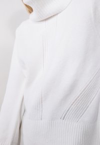 Maglione bianco a collo alto con texture a costine e sottili motivi verticali. Il tessuto appare morbido e accogliente.