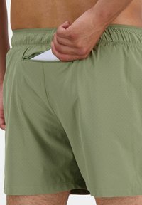 Grüne, durchlöcherte Badeshorts mit einem elastischen Bund und einer Gesäßtasche, in der ein Teil eines weißen Gegenstands teilweise eingelegt ist.