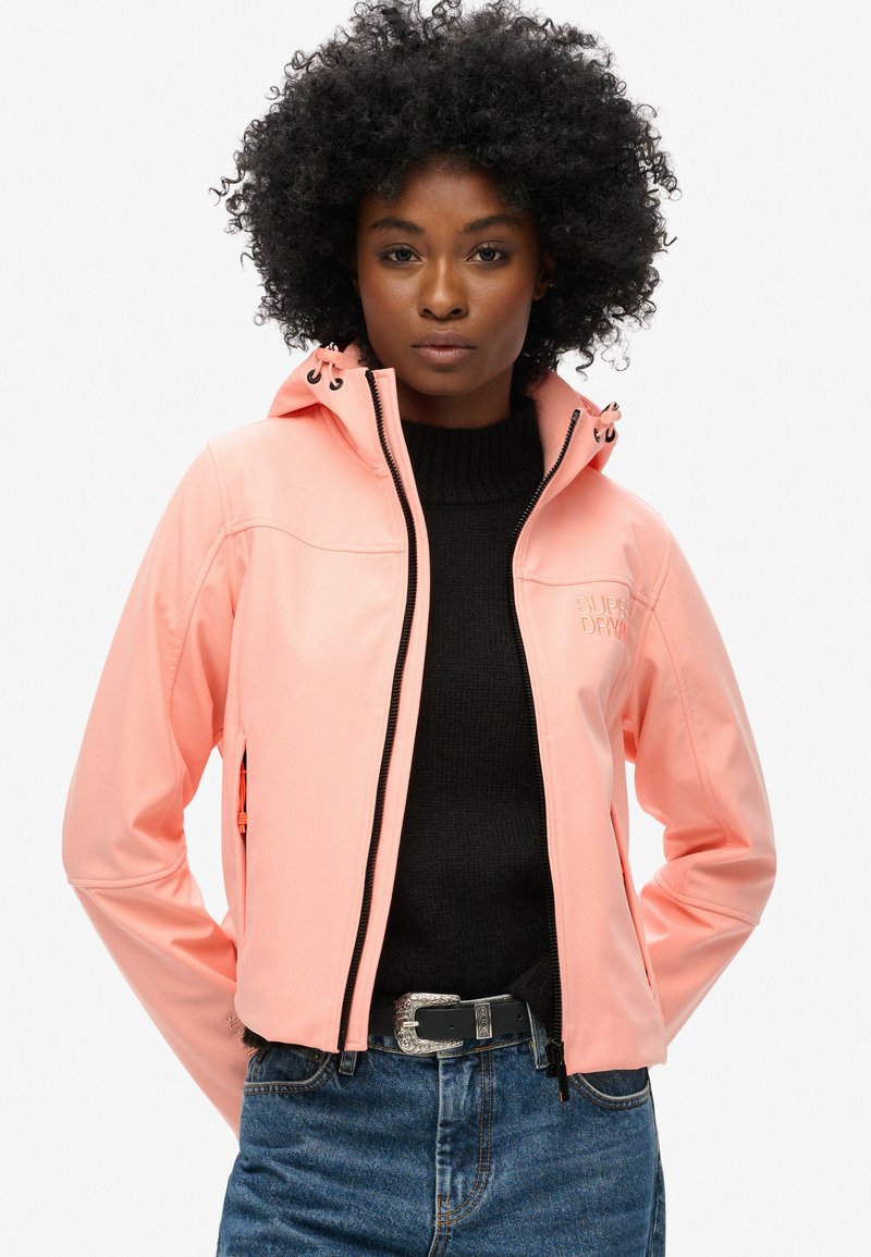 Korallpink softshell-jakke med hætte, fuld længde lynlås og logodetalje. Sammen med en sort sweater og denimjeans.