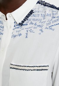 Vit skjorta med blå handskriven text, en ficka med flätad kant och en krage med en svart "be-strong"-tag.