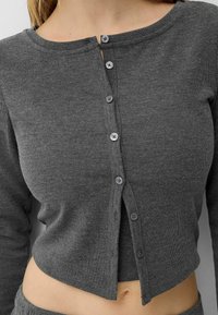 Grå, ribbad långärmad topp med rund halsringning och sex knäppningar framtill; kort design med råa kanter och slät textur.