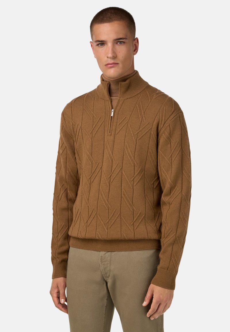 Brauner Strickpullover mit Reißverschlusskragen, der ein strukturiertes Blattmuster aufweist. Figurbetonter Schnitt, kombiniert mit hellen Hosen.