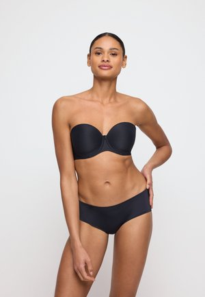 Mujer de pie con la mano en la cadera, llevando un sujetador negro sin tirantes y ropa interior negra a juego sobre un fondo liso y claro.