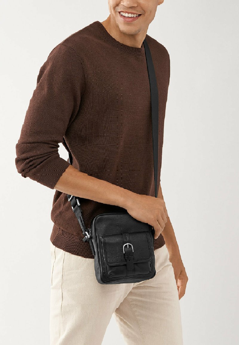Sac en cuir noir à porter en travers avec un rabat avant, une boucle en métal et une sangle réglable. Texture lisse et forme rectangulaire compacte.