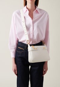 White leather crossbody bag with gold accents, rectangular shape, adjustable strap, and visible stitching detail. Worn with a pink shirt and dark jeans.