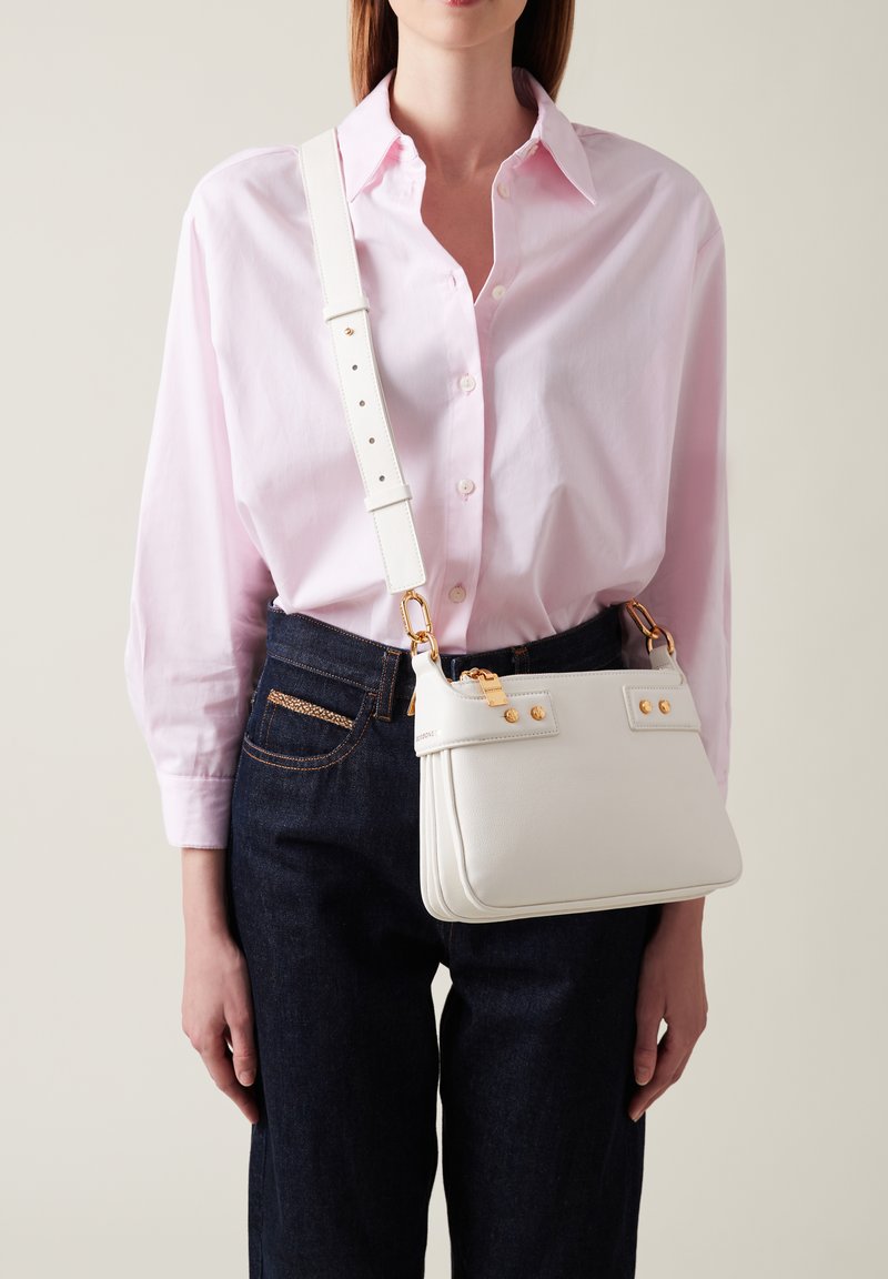 White leather crossbody bag with gold accents, rectangular shape, adjustable strap, and visible stitching detail. Worn with a pink shirt and dark jeans.